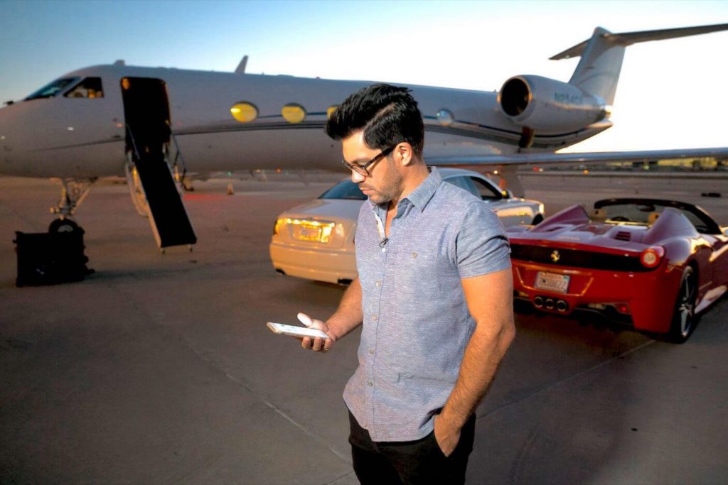 A man holding his cellphone behind two cars and a private plane in a runway