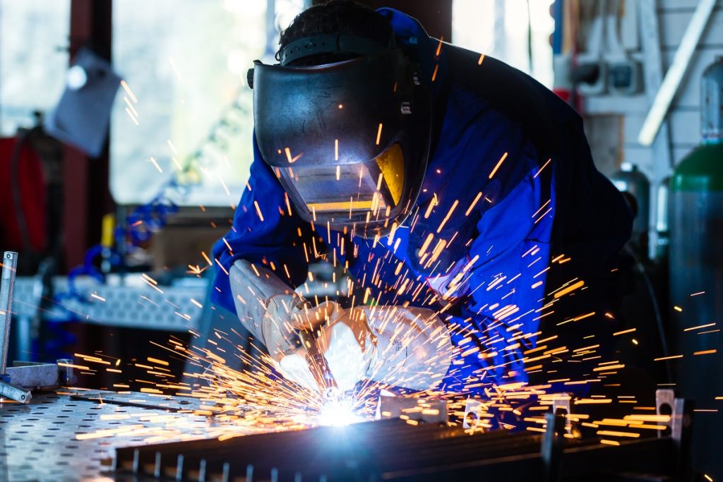 Welder bonding metal with welding device in workshop, lots of sparks to be seen, he wears welding goggles